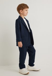 Mørkeblå fleece blazer med enkelt knap og sidelommer, parret med matchende bukser og hvide sneakers. Glat tekstur hele vejen igennem.