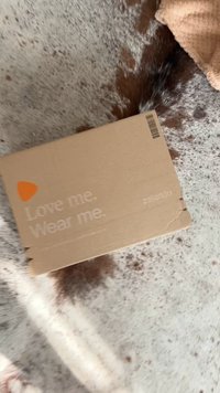 Caixa de cartão castanho com uma mensagem impressa, "Ame-me. Use-me." e logótipo. Cantos ligeiramente danificados, colocada sobre uma superfície texturada.