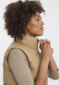 Veste rembourrée tan avec col montant, fermeture éclair et sans manches, portée par-dessus un haut à manches longues beige. Tissu texturé avec un motif matelassé.