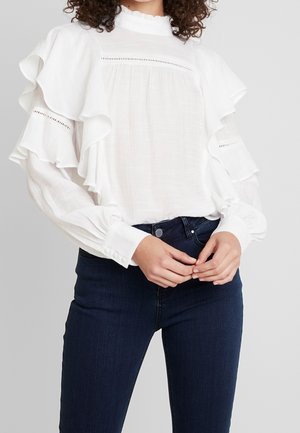 Vrouw draagt een witte blouse met lange mouwen met ruches en oogjesdetails gecombineerd met donkerblauwe spijkerbroek tegen een lichtgrijze achtergrond.