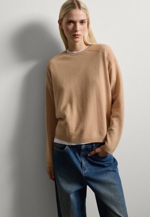 Suéter de punto beige con cuello de canalé y acentos en los hombros, llevado sobre una blusa blanca, combinado con vaqueros de denim azul holgados.