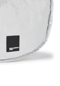 Sølv metallisk veske med en glatt, reflekterende overflate, med en svart kvadratisk etikett preget med "KARL LAGERFELD JEANS" nederst.