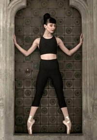 Top corto negro ajustado y leggings en una bailarina posando contra una pared de baldosas texturizadas, usando zapatos de punta beige. El cabello está recogido en un moño.