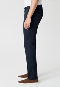Námořnické modré slim fit kalhoty vyrobené z bavlněné směsi, se zipem a bočními kapsami, nošené s hnědými běžeckými botami.