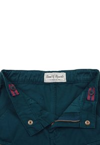 Vihreät orgaanisesta puuvillasta valmistetut housut, joissa on nappi- ja vetoketjusuljenta. Housuissa on vyötärölabel ja nappiin liittyvä raidallinen yksityiskohta.