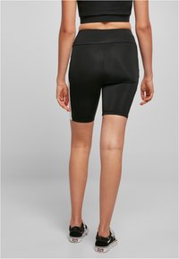 Osoba oblečená v čierno-bielych cyklistických šortkách, čiernom crop top a čierno-bielych teniskách, stojaca pred jednoduchým šedým pozadím.