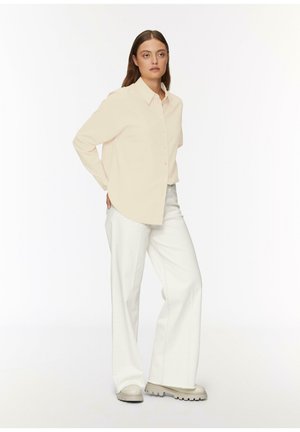 Femme aux longs cheveux bruns portant une chemise beige à boutons, un pantalon blanc à pattes d'éléphant et des chaussures à semelles épaisses beiges, se tenant les mains derrière le dos.