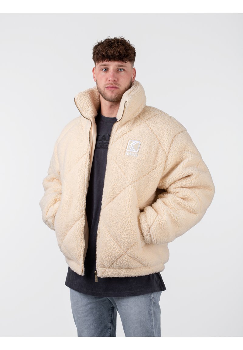 Chaqueta oversized acolchada beige, hecha de tela de felpa, con un cuello alto y cierre de cremallera, con un logo en la parte izquierda del pecho.