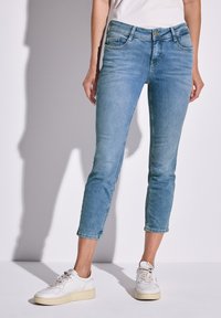 Jeans de denim azul claro con un diseño ajustado, longitud cortada y estilo de cinco bolsillos. Se llevan con zapatillas blancas sobre un fondo claro.