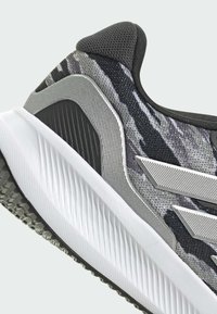 Athletischer Sneaker mit einem oberen Teil aus schwarzem und grauem Camouflage-Mesh, silbernen Akzenten, strukturierten Seitenpaneelen und einer weißen Gummisohle.
