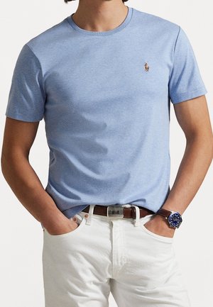 Mand iført lyseblå t-shirt med korte ærmer og rund hals, hvide bukser med brunt bælte og mørkeblå armbåndsur, hænderne i lommerne.