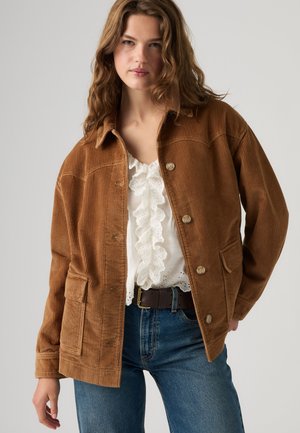 Donna con giacca di velluto a coste marrone con bottoni sopra una camicetta bianca arricciata e jeans blu, in piedi contro uno sfondo grigio uniforme.