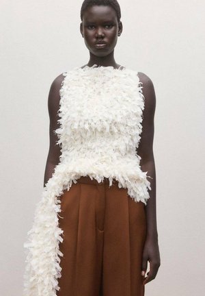 Persona de piel oscura con una blusa blanca sin mangas y texturizada, con una larga extensión de tela y pantalones marrones de talle alto, frente a un fondo liso.