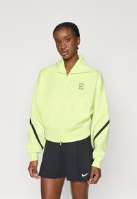 Sweatshirt zippé vert néon avec un col haut, accents de rayures noires sur les manches et un petit logo. Associé à une jupe de sport noire.