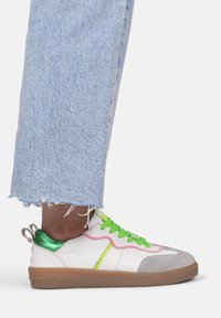 Weißer Sneaker mit grauem Wildleder-Zehenkappe, grünen reflektierenden Akzenten, leuchtend grünen Schnürsenkeln und pinken Nähten. Braune Gummisohle. Denim-Cropped-Hosen.