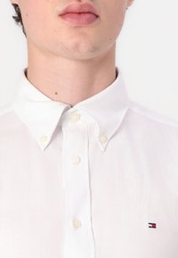 Cămașă albă cu nasturi dintr-o țesătură ușoară, având un guler clasic, placket frontal cu patru nasturi și un mic logo brodat pe piept.