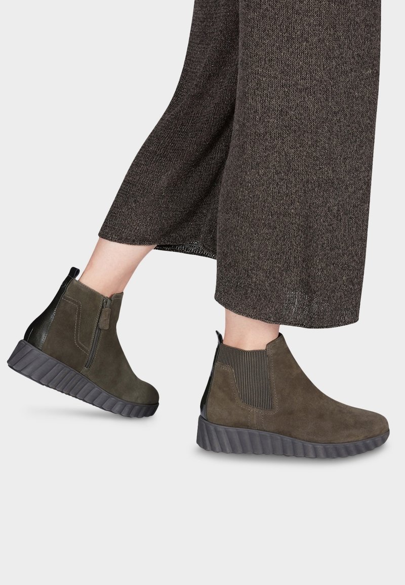 Stivaletti alla caviglia in camoscio verde con retro nero testurizzato, inserti elastici sui lati e suola spessa a coste. Indossati con pantaloni larghi marrone scuro.