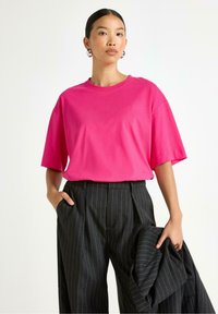 Jasnoróżowa bawełniana koszulka oversize z krótkimi rękawami, zestawiona z czarnymi spodniami w prążki o wysokim stanie. Model trzyma czarną kurtkę.