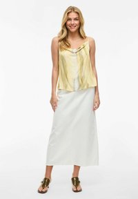 Top amarillo sin mangas con escote drapeado, combinado con una falda larga blanca. La modelo lleva sandalias marrones. Texturas suaves y diseño fluido.
