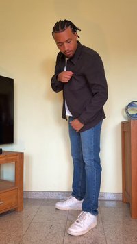 Casaco escuro e texturizado usado por cima de uma camisa branca, combinado com jeans azuis e ténis brancos. Postura de pé, a ajustar o bolso do casaco.