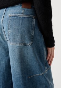 Unos vaqueros de mezclilla de color azul claro cuentan con un bolsillo trasero, una etiqueta de cuero y una silueta de pierna ancha, mostrando costuras visibles y una textura de tela suave.