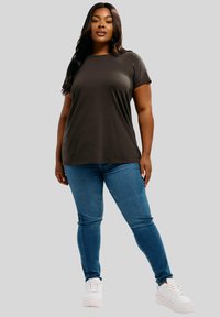 Donna che sta in piedi con sicurezza indossando una camicia grigio scuro a maniche corte, jeans skinny blu e sneakers bianche su uno sfondo neutro.