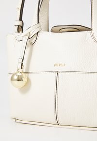 Weiße Ledertasche mit strukturierter Oberfläche, die ein goldenes Logo und einen runden goldenen Anhänger an der Seite aufweist. Schlichte Form mit zwei Griffen.