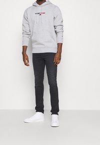 Szary bawełniany kaptur z kieszenią z przodu i logo "TOMMY JEANS" w czerwono-niebieskich barwach, zestawiony z ciemnymi dżinsami i białymi sneakersami.