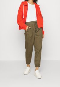 Chaqueta roja con capucha y cordones blancos, combinada con pantalones tapered en color verde oliva y zapatillas blancas. Diseño simple y funcional.