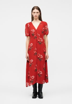 Femme portant une robe midi rouge à imprimé floral, manches bouffantes, boutons sur le devant, associée à des bottes noires montantes, debout devant un fond blanc.