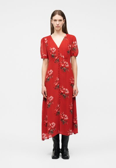Femme portant une robe midi rouge à imprimé floral, manches bouffantes, boutons sur le devant, associée à des bottes noires montantes, debout devant un fond blanc.