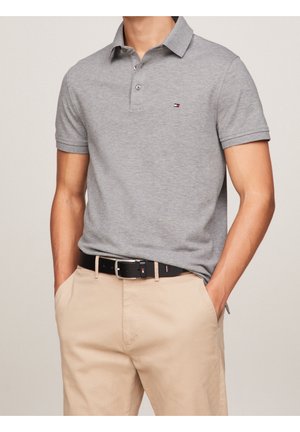 Polo gris à manches courtes, avec deux boutons au col et un petit logo sur la poitrine. Porté avec un pantalon beige et une ceinture noire.