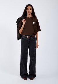 Camiseta de gran tamaño marrón con un gráfico de corazón blanco, combinada con jeans oscuros de pierna ancha y una chaqueta de denim llevada sobre el hombro.