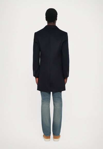 Marineblauer langer Mantel mit taillierter Passform, ausgestattet mit einem Einlass und zwei Knöpfen an den Ärmeln, kombiniert mit hellblauen Jeans und hellbraunen Schuhen.