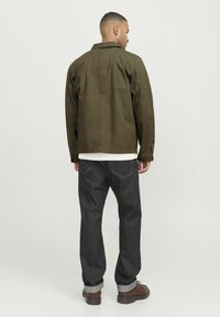 Giacca verde oliva con un design semplice, jeans in denim scuro e stivali in pelle marrone, con risvolti arrotolati e dettagli di cucitura nelle tasche posteriori.