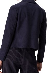 Vue arrière d'une personne portant un blazer bleu marine foncé et un pantalon noir à fines rayures sur fond blanc.