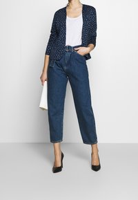 Blazer de patrón azul marino sobre camiseta blanca, jeans de mezclilla azul de talle alto, sosteniendo una toalla blanca y usando zapatos de tacón alto negros.