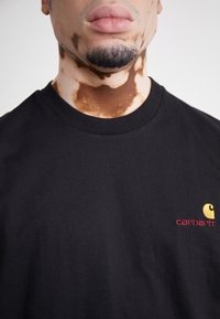Black cotton t-shirt with a crew neckline, featuring a small yellow and red Carhartt logo on the left chest. Soft texture, casual fit.