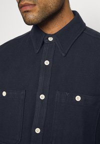 Mörkblå skjorta med knappar i strukturerad tyg, med spetstillverkad krage, två framfickor och beige knappar.