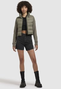 Jacket scurt, de culoare verde măsline, cu mâneci din tricot, purtat peste un top negru. Asortat cu pantaloni scurți negri cu talie înaltă și cizme negre cu șireturi.