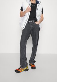 Vit pufferväst, svart t-shirt, mörkgrå jeans och flerfärgade sneakers (orange, gul, svart). Metallisk spänne-dekor.