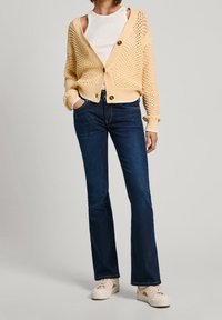 Person som bär en krämfärgad stickad kofta över en vit linne, mörkblå bootcut-jeans och beige sneakers, stående med händerna i fickorna.