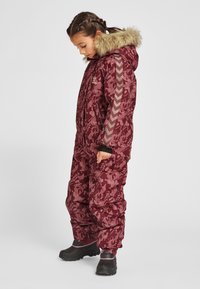 Burgundy winter snowsuit with floral pattern, faux fur-lined hood, gray chevron accents on sleeves, and zipper pockets. Black boots visible.
