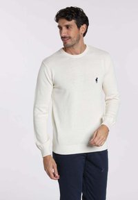 Pull en tricot de couleur crème avec un col rond, des poignets et un ourlet côtelés. Il présente un petit logo sombre sur la poitrine gauche.