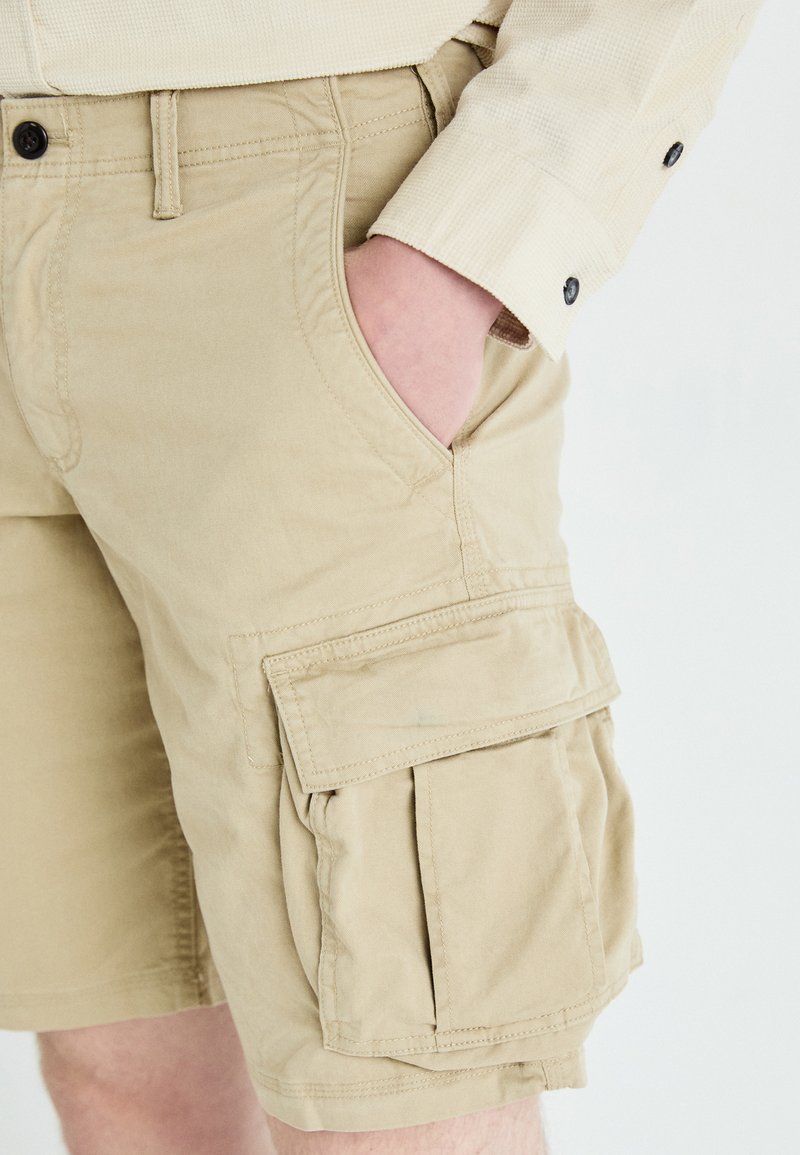 Shorts cargo beiges en coton, dotés de poches avant et de grandes poches latérales avec rabats, tissu texturé et détails de couture visibles.