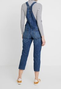 Denimoverall i mörkblå tvätt med justerbara axelremmar, två bakfickor och uppvikt anklar, kombinerat med en randig långärmad tröja.