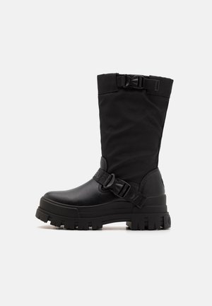 Botas pretas de cano médio feitas de material sintético, com um cabedal texturizado, tira com fivela e sola em borracha espessa com sulcos profundos.