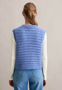 Jasnoniebieski, dziany kamizelka o teksturowanym wzorze i ściągaczu, noszona na białej bluzce z długim rękawem, w połączeniu z niebieskimi dżinsami.
