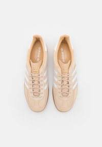 Bruna mockasinsneakers med vita läderremsor, rund tå, platt snördesign och vadderad krage; har guldlogotyp på tungan.