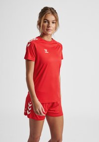 Röd idrottströja och shorts-set, tillverkat av lättviktsmaterial. Har korta ärmar, rund hals och vita chevrondetaljer på axlarna.